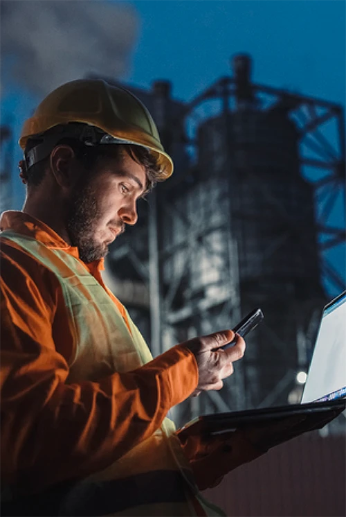 Lone worker working at night outside a factory, using smartphone and portable laptop device