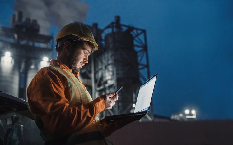 Lone worker working at night outside a factory, using smartphone and portable laptop device